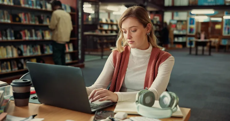 Mloda studentka pisze na laptopie przy stole w bibliotece uczelni, obok kubek kawy i sluchawki